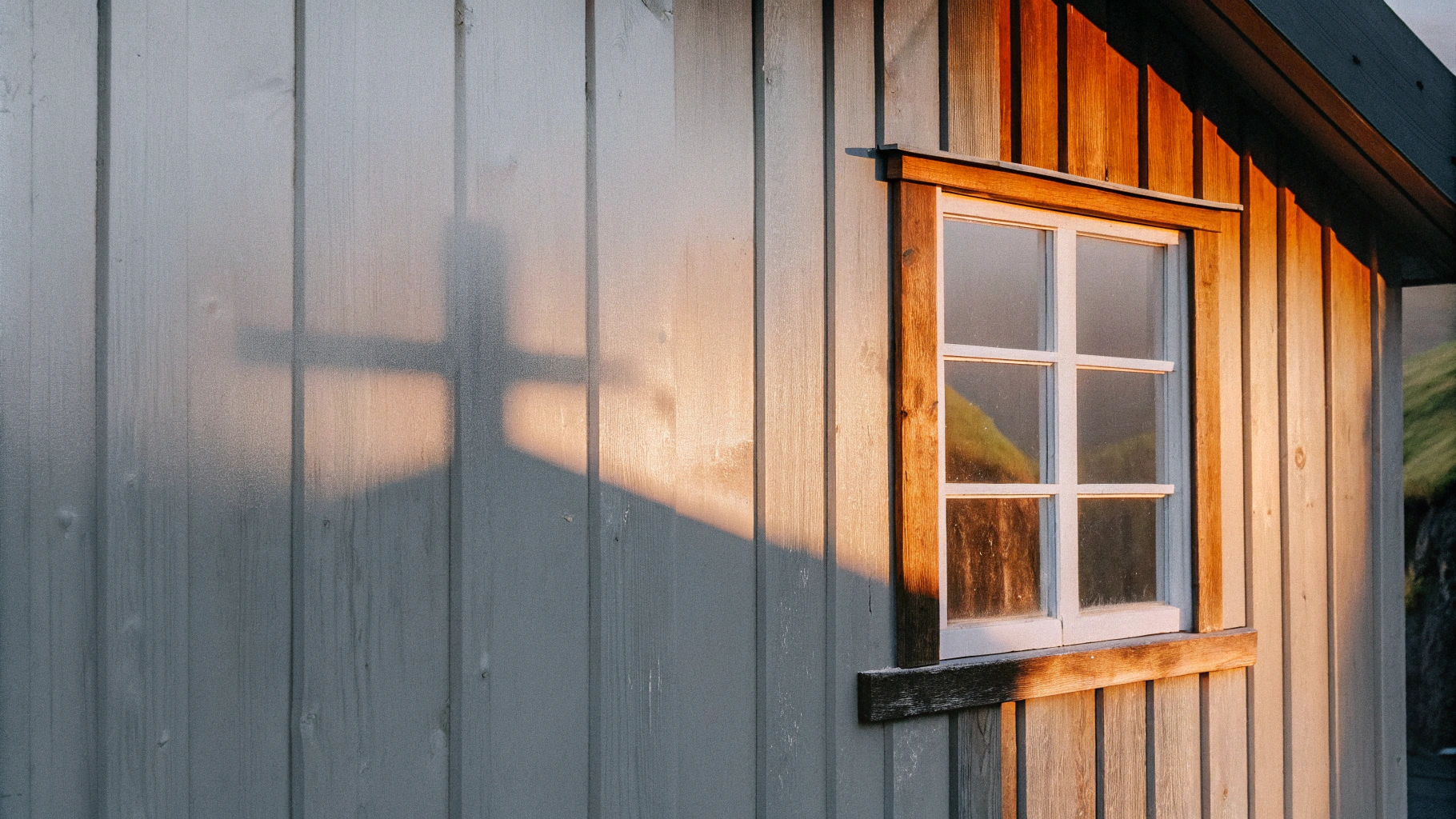 AI generated image by Red Panda AI: Sunlight breaks through the fog and casts a warm, golden glow into a small, weathered wooden cottage in the Faroe Islands. To one side, a single window with cross-shaped panes lets in the light, projecting the shadow of the cross onto the opposite wall. In the center of the scene, this shadow rests prominently on the wall, like a sacred symbol. Outside, close to the window, a green, moss-covered cliff face is visible, enhancing the still, timeless atmosphere of the quiet refuge.