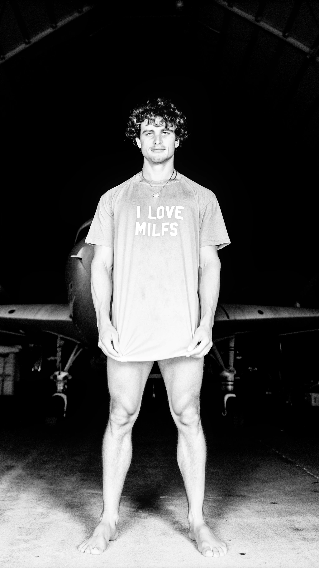 AI generated image by Red Panda AI: A young fit muscular handsome tall boy with curly hair standing infront of a F35 lightning 2 wearing an oversized tshirt with words written "I love milfs" on it , in a shed with a lot of  Machinery in background, dark background , F35 