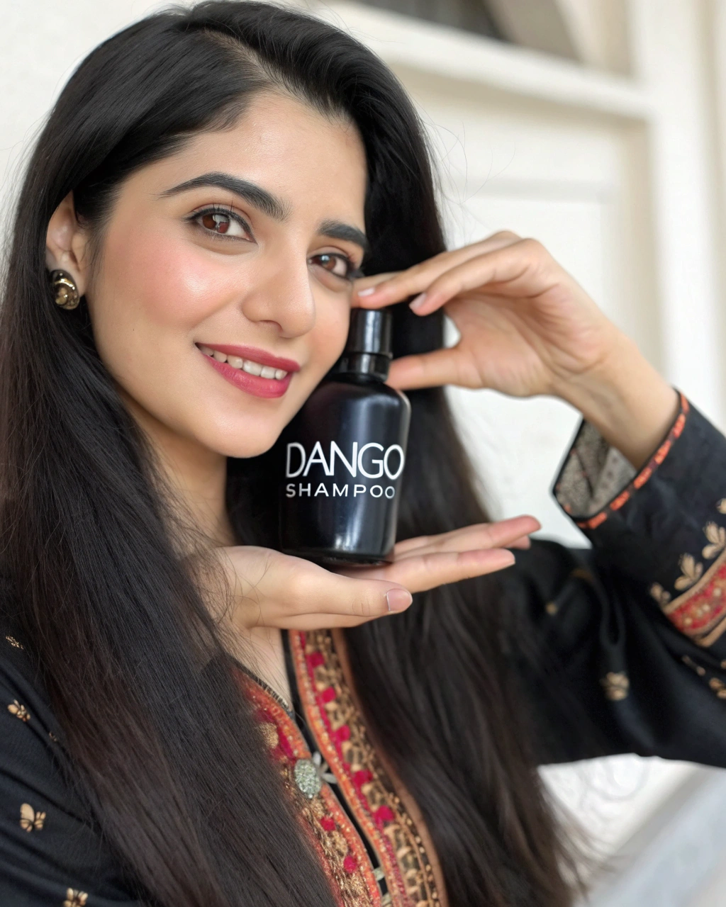 AI generated image by Red Panda AI: A beautiful Pakistani model closeup having black hair and wearing kamiz for a brand showing off a shampoo small black bottle no longer than her hand with "DANGO Shampoo" written on it with a white label.