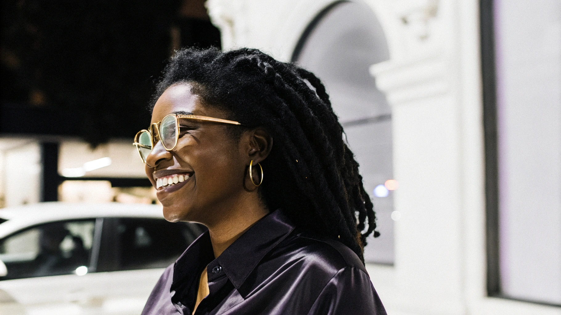 AI generated image by Red Panda AI: A black woman with natural curly hair in nagô braids, wearing gold-rimmed glasses and a purple silk blouse, smiling spontaneously. Blurred background with a neon-lit store in shades of turquoise and lilac. 35mm lens, portrait, natural night lighting, vibrant, urban style, fashion, beauty.