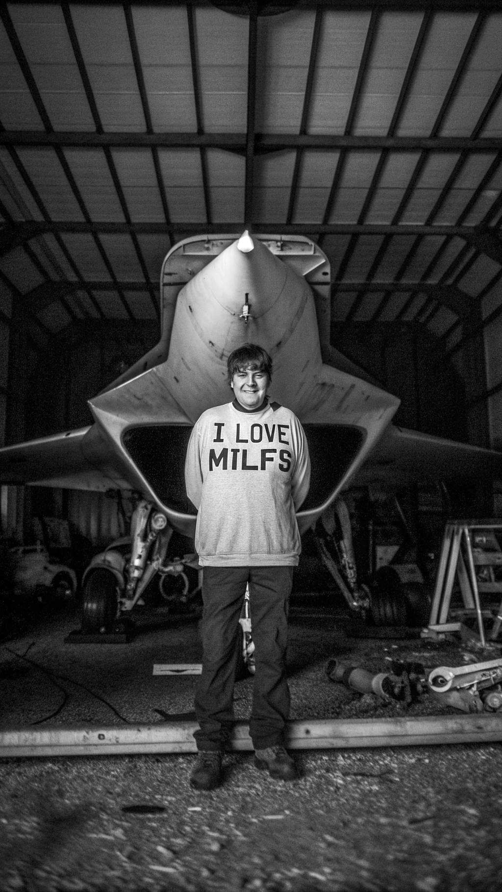 AI generated image by Red Panda AI: A young man standing infront of a F35 lightning 2 wearing an oversized tshirt with words written "I love milfs" on it , in a shed with a lot of  Machinery in background, dark background , F35 