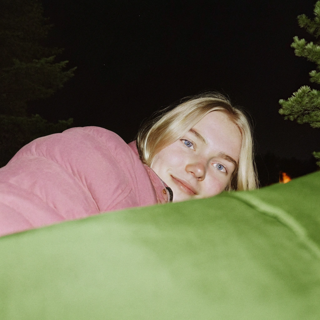 AI generated image by Red Panda AI: A raw photo taken from a selfie POV at night, showing a beautiful 18-year-old blonde woman with pale skin and stunning azure blue eyes. She’s dressed warmly, with a subtle, gentle smile. The background shows the dark night sky, the glow of a nearby campfire casting soft light on her face, and faint shadows of trees surrounding the campsite. High-resolution, capturing fine details of her skin and her bright blue eyes, while the warm firelight creates a peaceful, cozy ambiance in the outdoor setting.