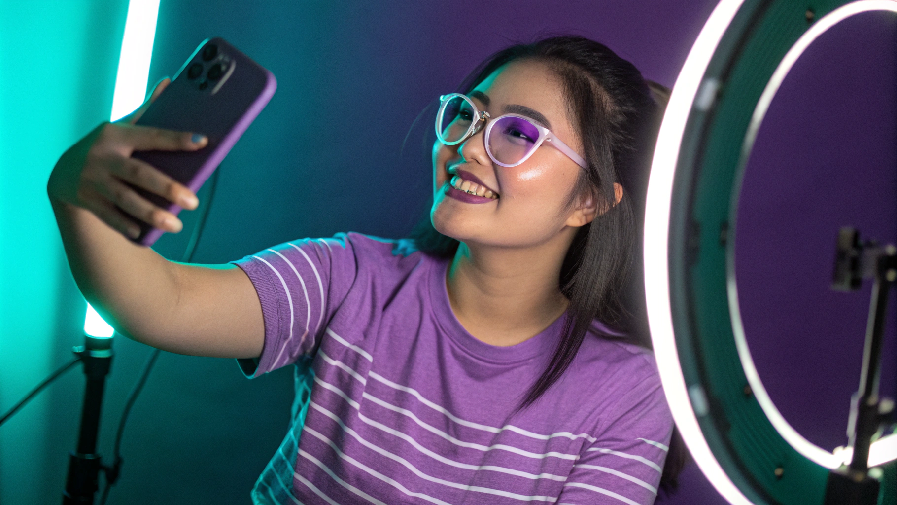 AI generated image by Red Panda AI: Uma fotografia de uma jovem descendente de japoneses tirando uma selfie em um estúdio iluminado com luzes neon roxas e turquesa, criando uma atmosfera vibrante e moderna. Ela está em frente a um ring light de LED, que destaca suas feições. A jovem veste uma camiseta lilás com finas listras brancas horizontais e usa óculos com lentes lilases. Sua maquiagem é marcante, com sombras de olhos em um tom de azul turquesa que realçam seu olhar. A imagem é um retrato realista, capturando detalhes de forma precisa e com uma iluminação que valoriza sua expressão alegre e estilo.