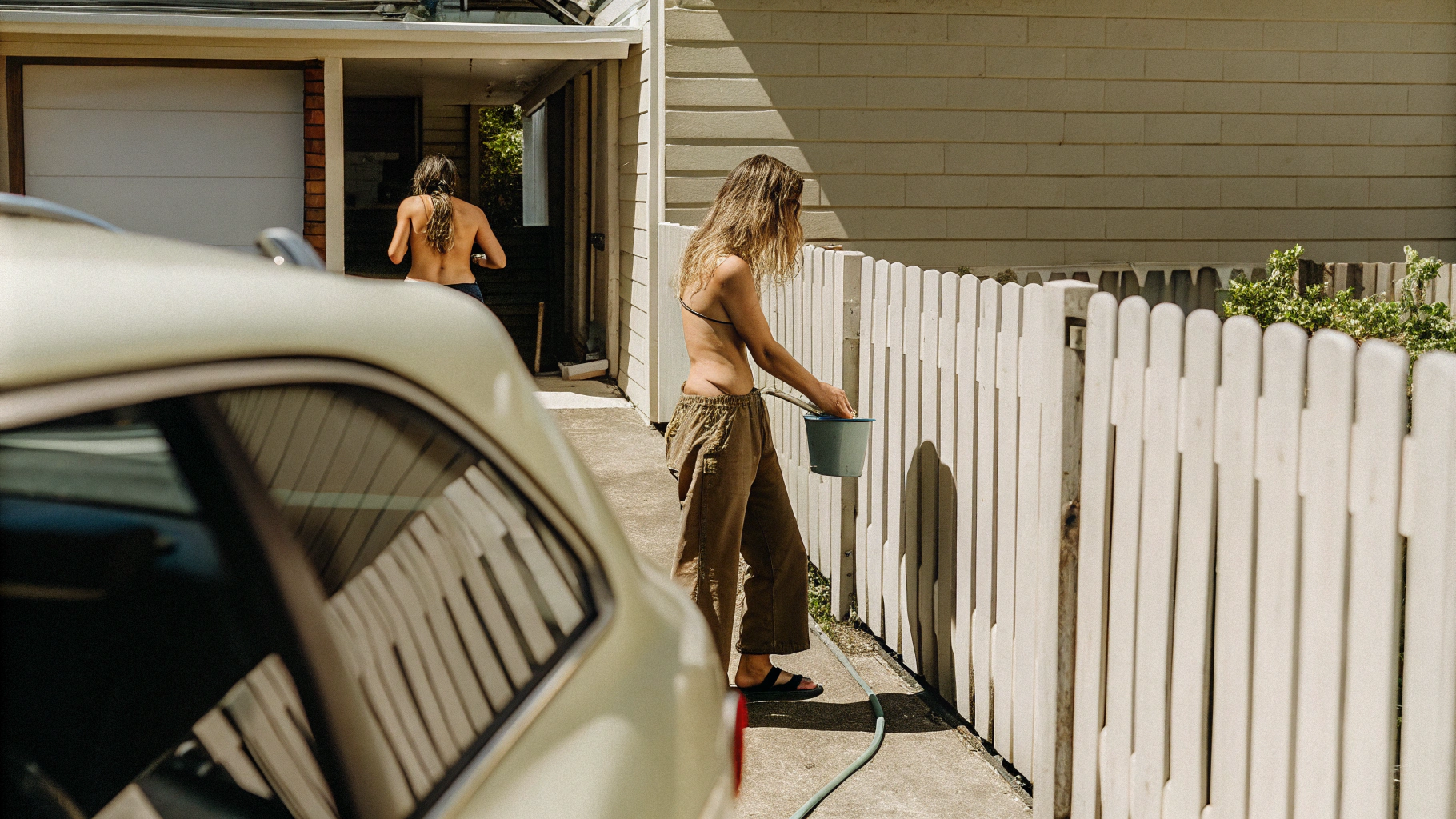 AI generated image by Red Panda AI: Dslr shot of a scandinavian woman i casual clothing, in her 30s is washing her car mid day with out a shirt on, and the neighbours is looking over the fences and he ges without being seen