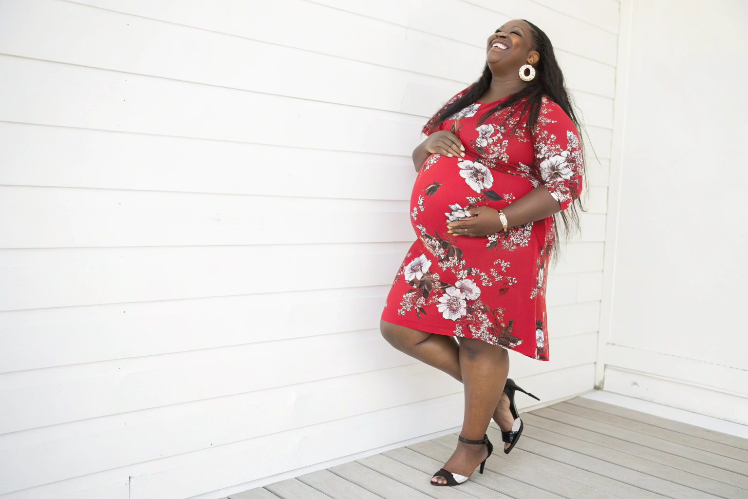AI generated image by FLUX.1-schnell: A pregnant black woman, plus size, with long hair, smiling, dressed in a red floral print dress, heels, and diamond earrings, standing against a white background.

