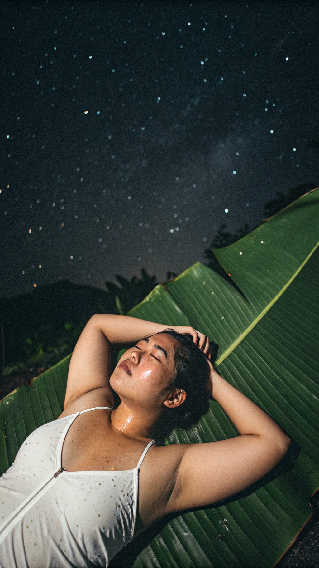AI generated image by Red Panda AI: tampilkan foto menyeluruh seorang Wanita Indonesia, muda, imut, agak gemuk, berlumuran keringat, dada membusung, ekspresi kecapekan, ia sedang berbaring di luar ruangan beralaskan daun pisang, pada malam hari yang gelap, pengambilan gambar diambil dari atas. Tank top putih dengan tali tipis di bahu, arms behind head. efek disposable.