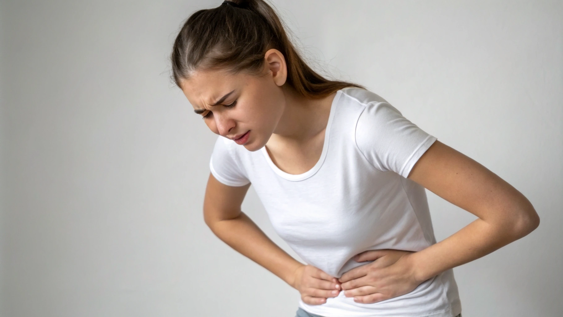 AI generated image by Red Panda AI: A young woman shown from the shoulders to the hips, focusing on her torso and hands pressing against her abdomen, conveying a sense of pain or discomfort. She is holding her stomach with both hands, showing discomfort from abdominal pain. The background is plain and minimal, emphasizing her expression and posture. She isThe lighting is soft and neutral, creating a clinical, health-related atmosphere.


