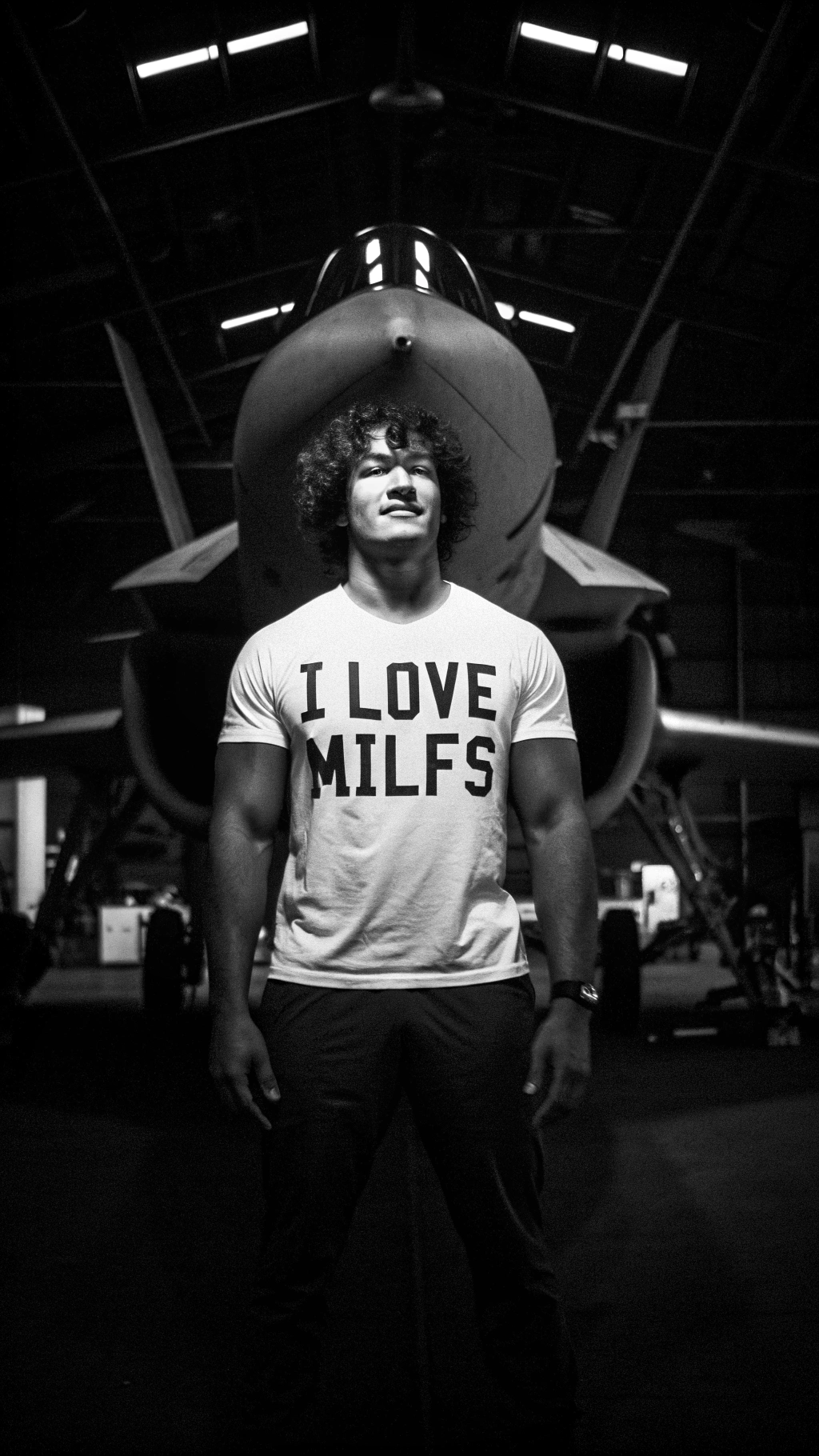 AI generated image by Red Panda AI: A young fit muscular handsome tall boy with curly hair standing infront of a F35 lightning 2 wearing an oversized tshirt with words written "I love milfs" on it , in a shed with a lot of  Machinery in background, dark background , F35 in background, F35 , ultra futuristic 