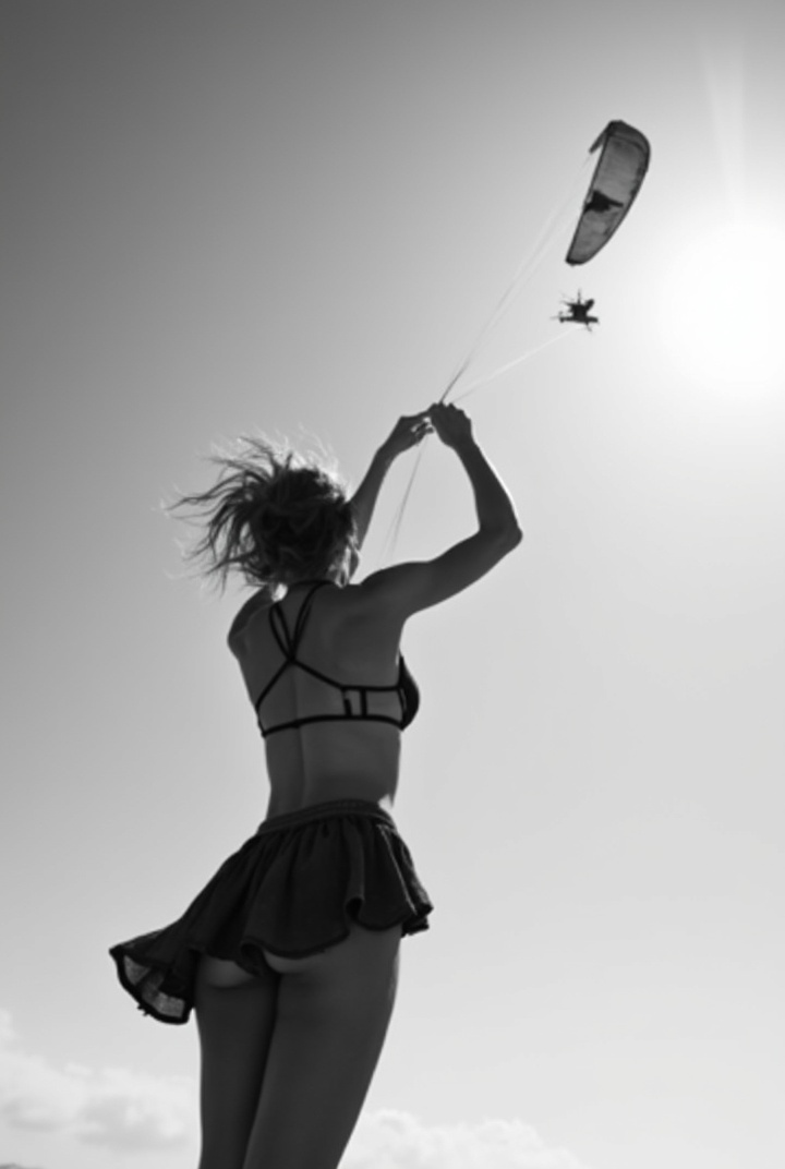 AI generated image by FLUX-Realism-Lora: Black and white photo of bare back woman in short skirt from behind swinging her bra above. a paraglider flying above her. Sky background. Backlight