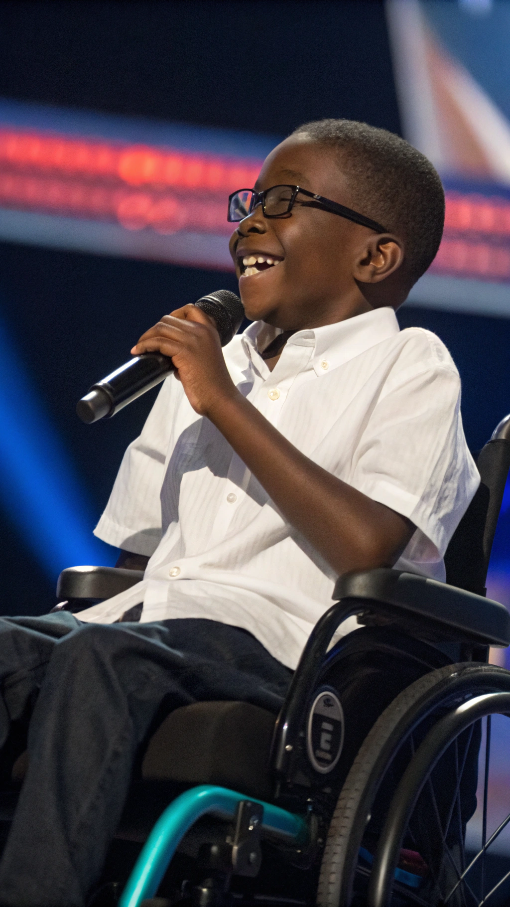 AI generated image by Red Panda AI: "Un niño africano ciego, de piel oscura y cabello corto, sentado en una silla de ruedas moderna. Sostiene un micrófono con firmeza en su mano derecha, mientras transmite confianza y alegría. Lleva una camisa blanca de algodón y pantalones oscuros, con un diseño limpio y bien cuidado. Sus gafas oscuras reflejan su ceguera, y su sonrisa ilumina la escena. El fondo es de America's Got Talent, con luces suaves resaltando los detalles de su rostro y la textura de su piel., creando una atmósfera inclusiva y emotiva. Renderizado en 8K, con iluminación cinematográfica y un enfoque que resalta la fuerza y resiliencia del niño. simula que el niño esta cantando 