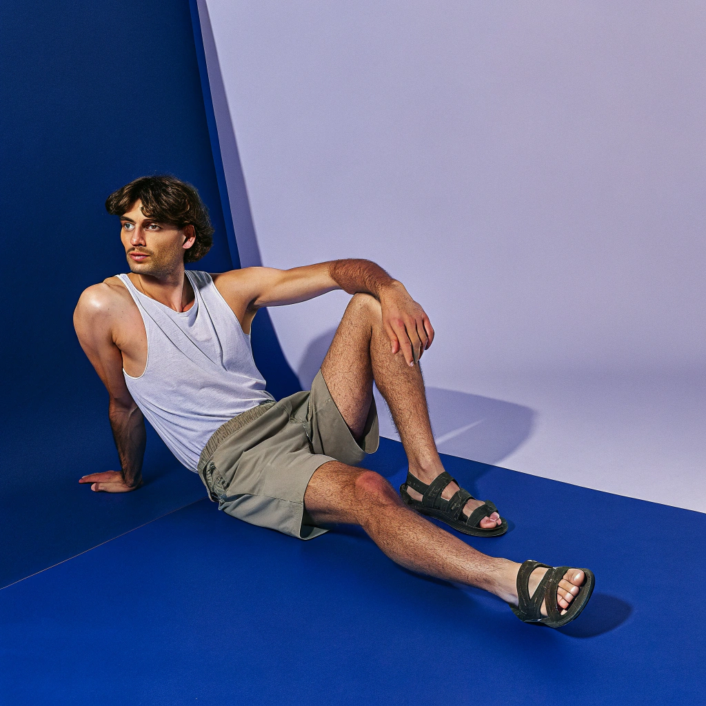 AI generated image by Red Panda AI: Uma cena ultra realista de um modelo masculino, corpo atlético bronzeado do sol, cabelos médios e castanhos com corte de cabelo estilo surfista. Posando para foto com camiseta regata branca estilo tank, calção curto de linho na cor caqui e sandálias com tiras cruzadas verde militar. IMG_1025.HEIC