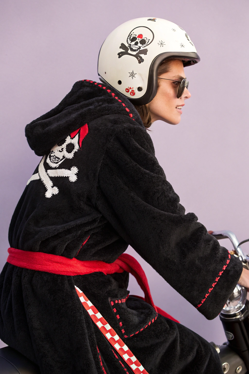 AI generated image by Red Panda AI: side portrait editorial fashion studio portrait of cute skull racing on a motor bike and wearing minimal styled black plush bathrobe Versace like with cute white pirate skull pattern and a red bathrobe belt and a cute helmet, pastel purple background, cinematography and photoshot by Wes Anderson and David LaChapelle