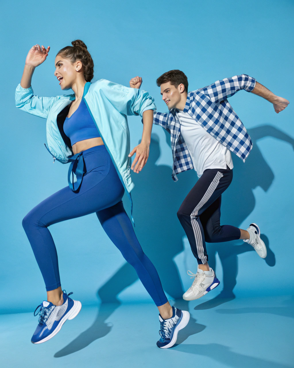 AI generated image by Red Panda AI: Two young adults, one male and one female, in a dynamic, energetic pose on a bright blue background. The female is in the foreground, dressed in sporty attire with a tight-fitting, bright blue top, matching leggings, and a light jacket tied around her waist. She is wearing stylish sneakers, and her hair is tied back. The male in the background wears a blue and white checkered shirt, dark pants, and matching sneakers. Both are mid-motion, with expressive body language that conveys movement and athletic energy. The lighting is clear and bright, emphasizing the vibrancy of their clothing and the modern, sporty aesthetic.