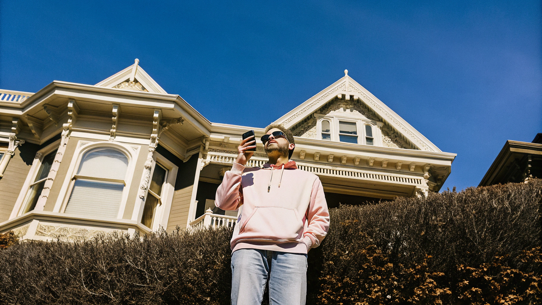 AI generated image by Red Panda AI: A 45 years old man wearing a pink hoodie and jeans. He is standing in front of a Victorian house in San Francisco. The picture has a vibrant day light in spring time and was taken with a iPhone 15 pro.